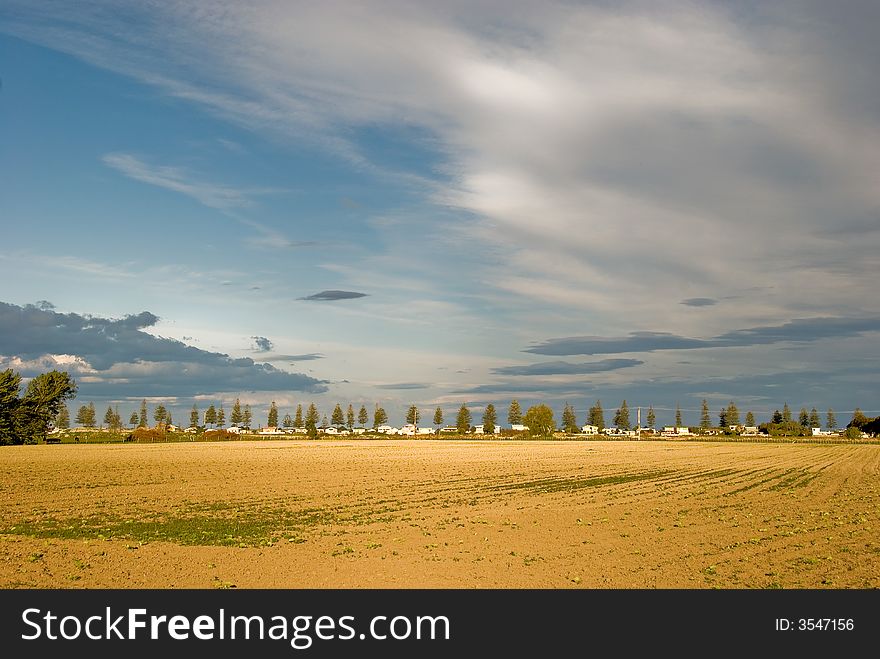 View across ploughed/plowed field to tree line and houses with big sky beyond. View across ploughed/plowed field to tree line and houses with big sky beyond