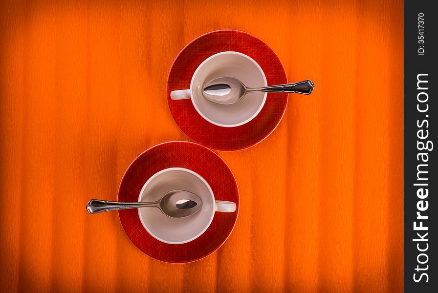 Two white empty cups with tea spoons, on red plates over orange color background, view from above