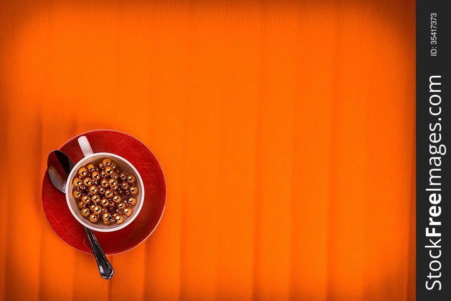 One lovely cup with golden beads in it , little fork and spoon by sides. Over textured bright orange color background. One lovely cup with golden beads in it , little fork and spoon by sides. Over textured bright orange color background.