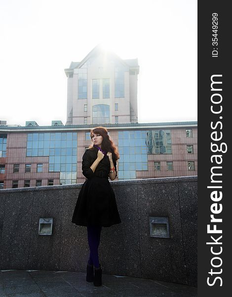 Young Woman Standing In Black Coat On Urban Background