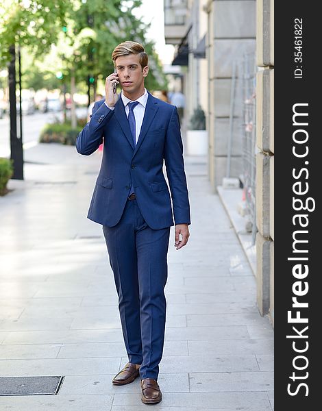 Portrait of an attractive young businessman on the phone in urban background, wearing blue suit and tie. Blonde hair