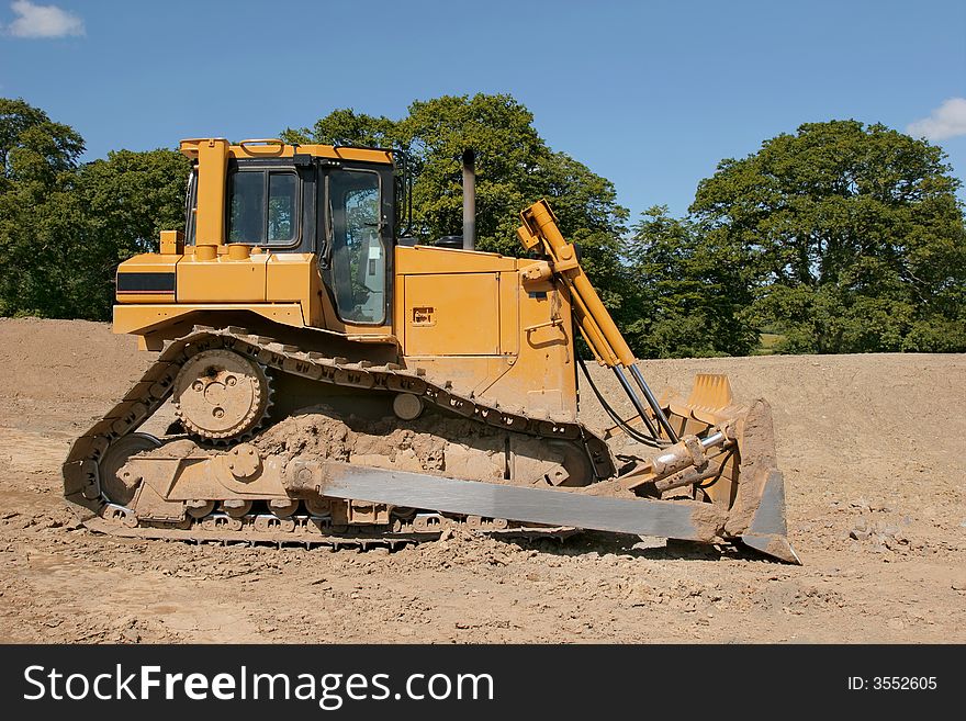 Yellow bulldozer (side view) standing idle on rough earth with trees and a blue sky to the rear.