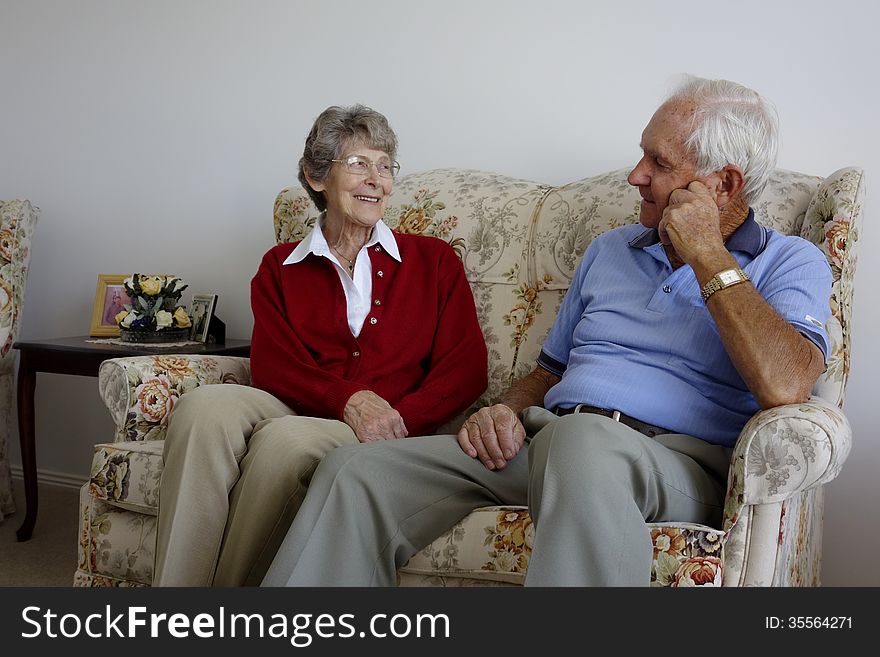 An elderly couple sit and relax on a couch as engage in conversation and enjoy each others company.