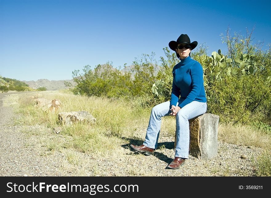 A woman wearing a cowboy hat sitting on a large cut rock by the side of the road. A woman wearing a cowboy hat sitting on a large cut rock by the side of the road.