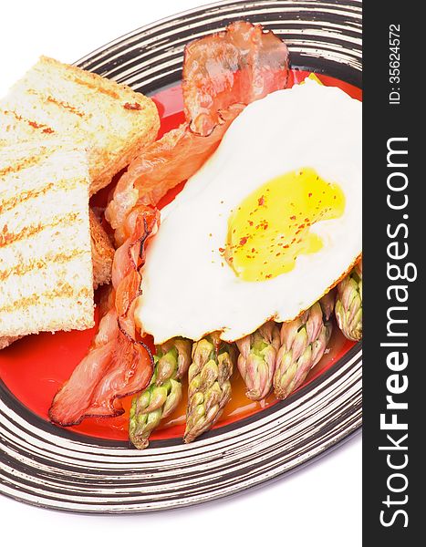 Tasty Breakfast with Fried Egg Sunny Side Up on Delicious Asparagus Sprouts, Roasted Bacon and Toasts on Red Striped Plate closeup on white background
