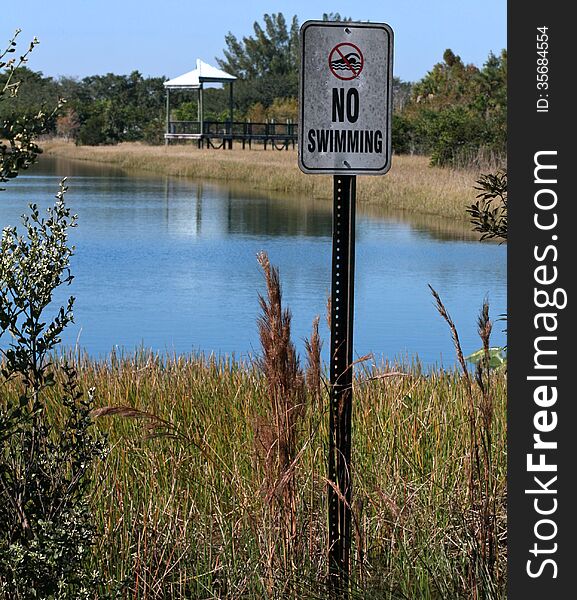 No Swimming sign beside lake at park with picnic area in background. No Swimming sign beside lake at park with picnic area in background