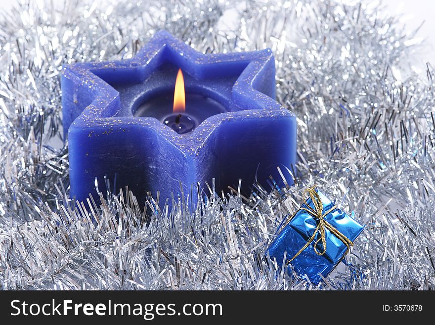 A deep blue six sided christmas candle nested in silver tinsel with blue giftbox.