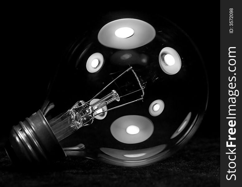 Black and white close up of a clear light bulb with white lights shining on it, reflecting off the glass of the bulb. Set off against a black background.