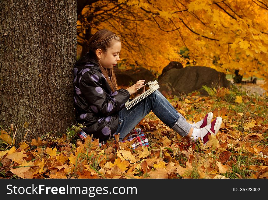 Pretty girl sitting under a tree and busy playing on the tablet on the background of the autumn landscape. Pretty girl sitting under a tree and busy playing on the tablet on the background of the autumn landscape