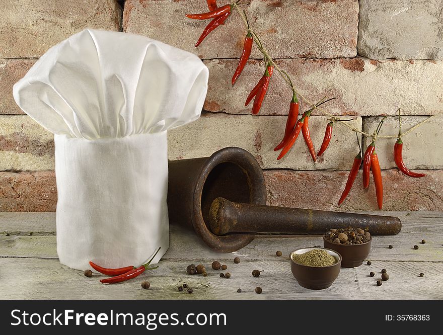 White toque with old mortar and pestle, whole chili peppers and peppercorn on brick wall. White toque with old mortar and pestle, whole chili peppers and peppercorn on brick wall