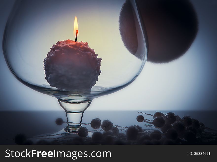 Small white candle burning and melting like a snowball inside big glass goblet. Over grey tint background with one big bulb and many small bubbles on the table. Small white candle burning and melting like a snowball inside big glass goblet. Over grey tint background with one big bulb and many small bubbles on the table.