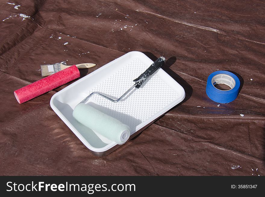 Painting supplies on a protective paint tarp that include a paint tray, rollers, brush and tape. Painting supplies on a protective paint tarp that include a paint tray, rollers, brush and tape.