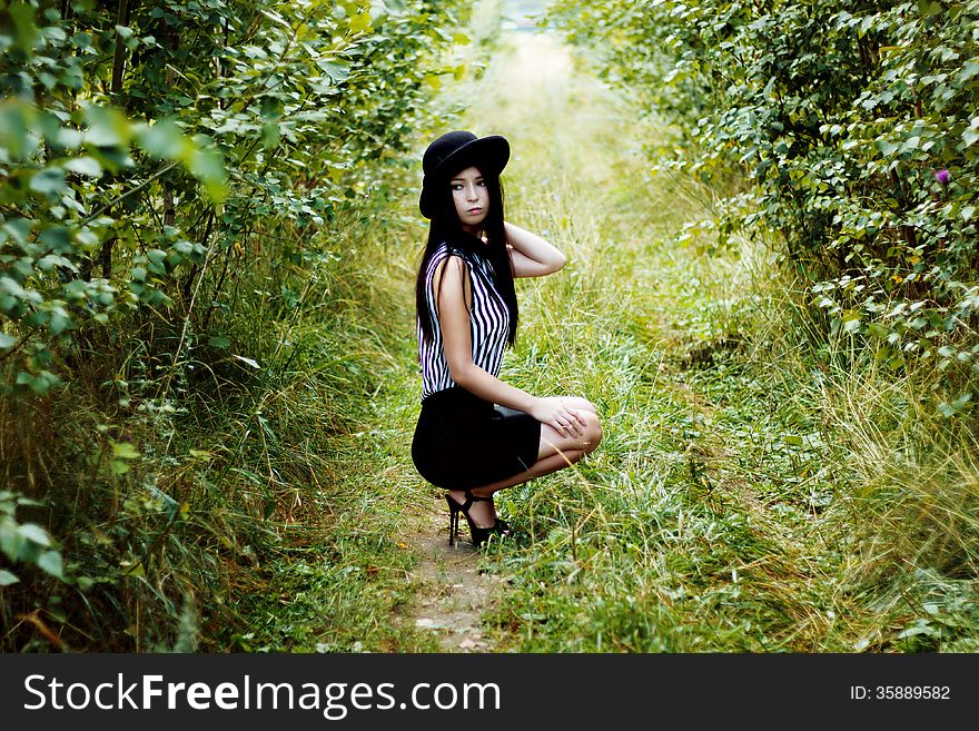 Fashionable woman in black hat sitting in green forest