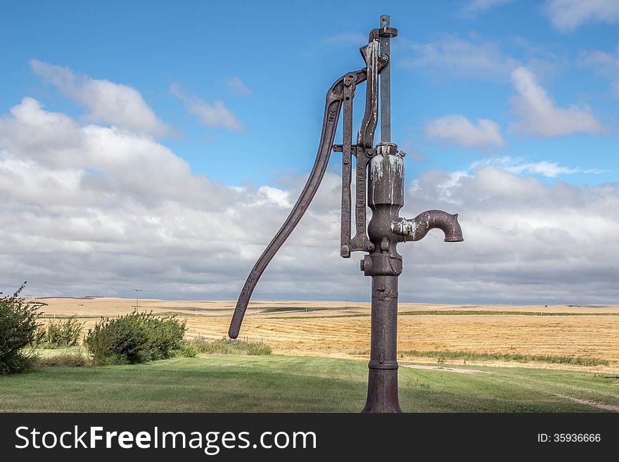 Old rusty antique water pump sitting in a field of golden wheat and green grass and a bright blue cloud filled sky. Old rusty antique water pump sitting in a field of golden wheat and green grass and a bright blue cloud filled sky