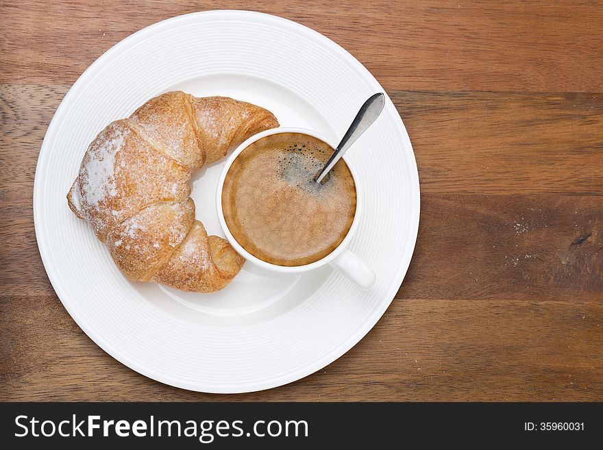 Fresh croissant and cup of coffee on a plate on wooden background, top view, close-up. Fresh croissant and cup of coffee on a plate on wooden background, top view, close-up
