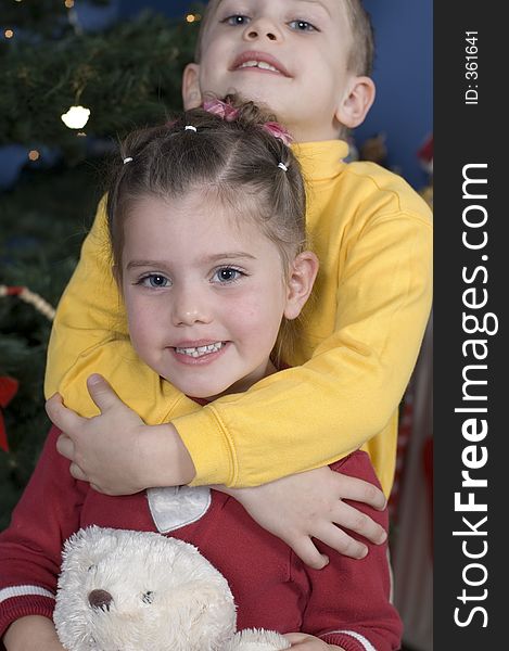 In front of a Christmas tree, boy stands behind his sister hugging her neck, the sister is holding a teddy bear. In front of a Christmas tree, boy stands behind his sister hugging her neck, the sister is holding a teddy bear