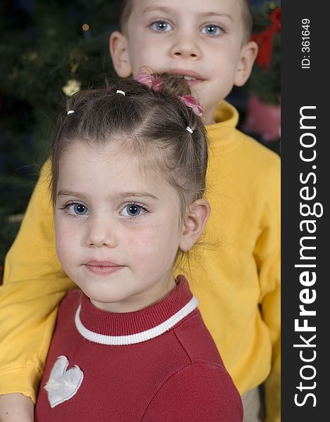 In front of a Christmas tree, boy stands behind his sister with his arm around her while she turns sideways and looks into the camera. In front of a Christmas tree, boy stands behind his sister with his arm around her while she turns sideways and looks into the camera