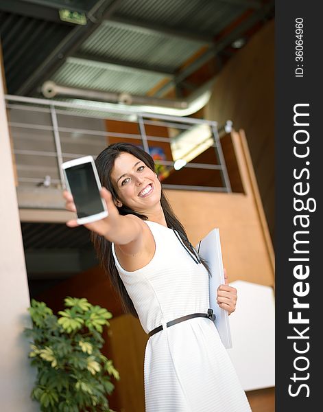 Modern professional businesswoman showing cellphone screen for inviting to call. Business woman portrait inside corporate building. Modern professional businesswoman showing cellphone screen for inviting to call. Business woman portrait inside corporate building.