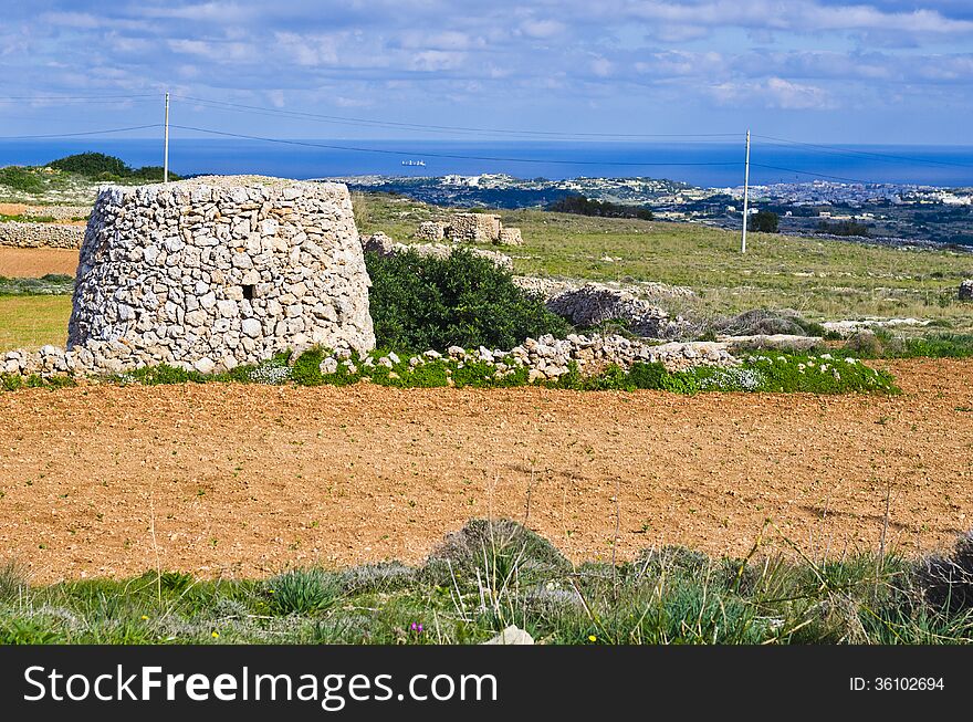 A corbel led stone hut and a cultivated field with crops in the maltese countryside. In the background is the island of Gozo visible surrounded by clear blue Mediterranean sea water. A corbel led stone hut and a cultivated field with crops in the maltese countryside. In the background is the island of Gozo visible surrounded by clear blue Mediterranean sea water.