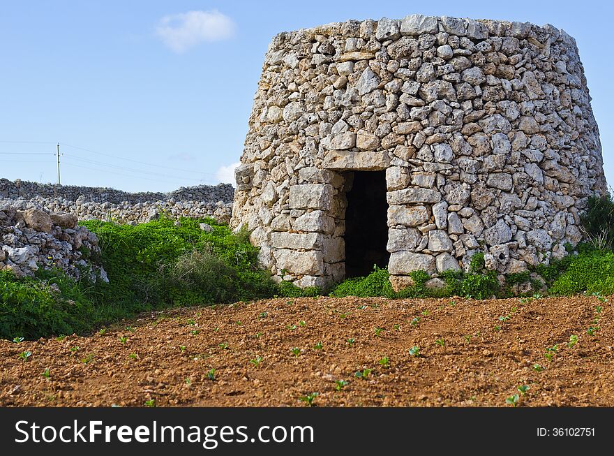 A corbel led stone hut and a cultivated field with crops in the maltese countryside. In the background is the island of Gozo visible surrounded by clear blue Mediterranean sea water. A corbel led stone hut and a cultivated field with crops in the maltese countryside. In the background is the island of Gozo visible surrounded by clear blue Mediterranean sea water.