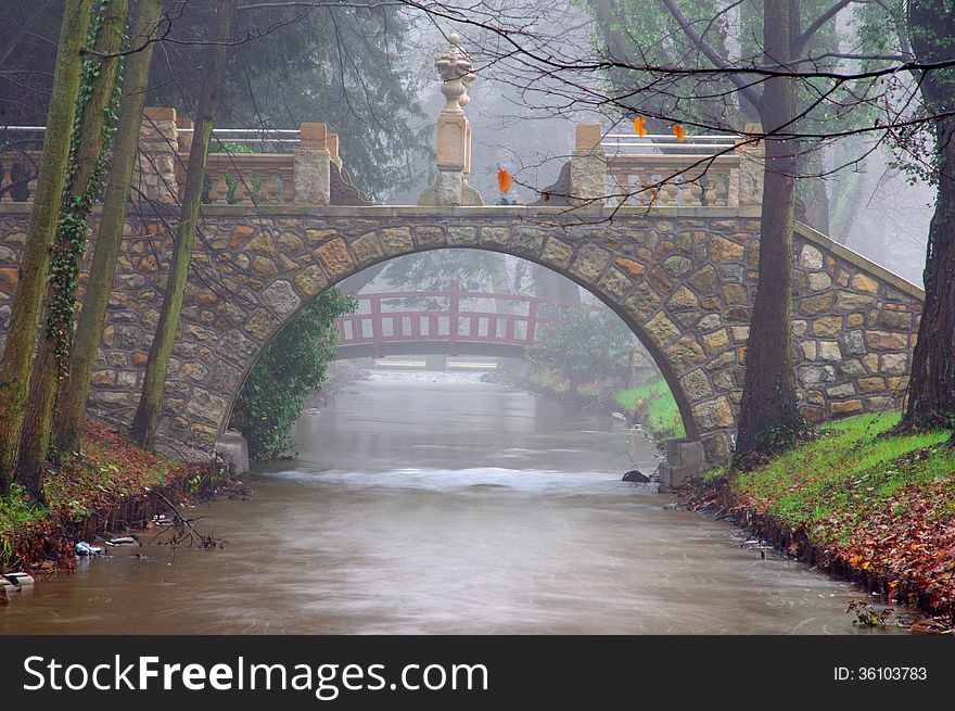 The photograph shows a small river flowing through the park. Passed over the river is a stone bridge for pedestrians. The photograph shows a small river flowing through the park. Passed over the river is a stone bridge for pedestrians.