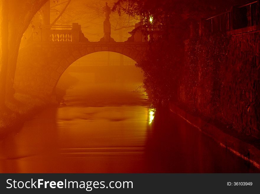 The photograph shows a small river over which passed the arched stone bridge. It is a misty night. Glow illuminates the dark area on the left side of the cadres of electric lamps. The photograph shows a small river over which passed the arched stone bridge. It is a misty night. Glow illuminates the dark area on the left side of the cadres of electric lamps.