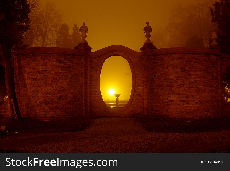 The photograph shows the park at night. Light illuminates the darkness of the park lamps. Above ground fog rises. In the scene we see the brick, corrugated wall with oval holes.