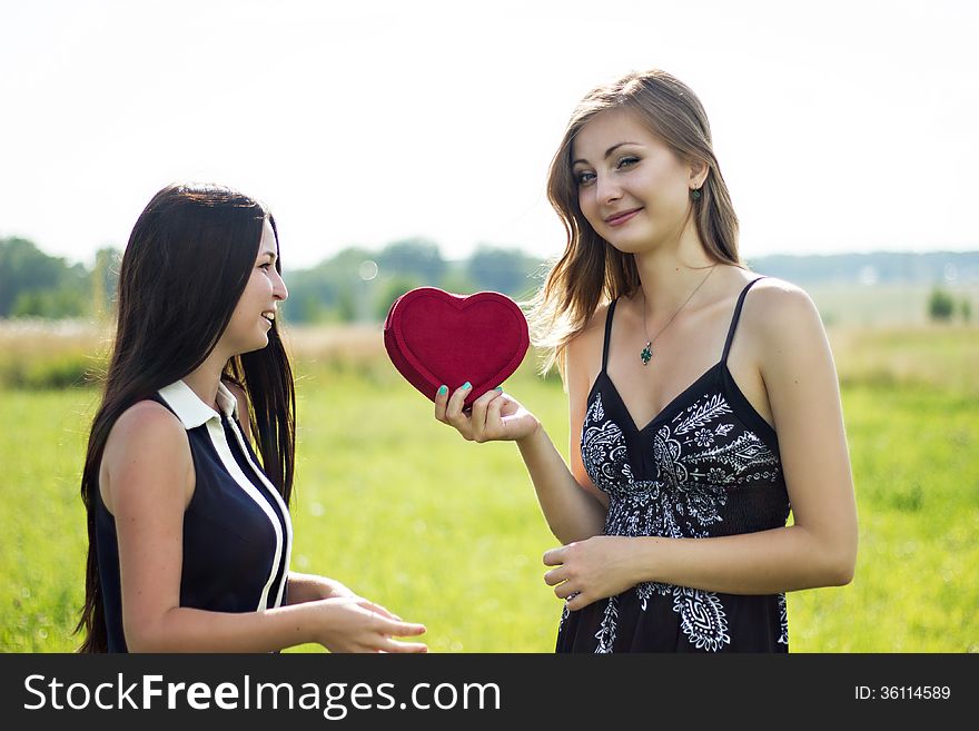 Two pretty women in love with red heart in sunshine summer field