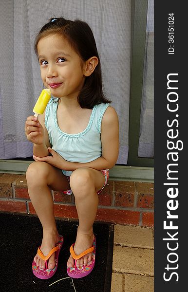 A young Asian girl sits on an outdoor step and grins as she takes a bite of her very cold icy-pole. A young Asian girl sits on an outdoor step and grins as she takes a bite of her very cold icy-pole.