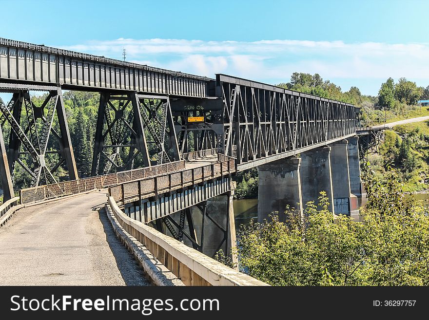 Black steel car bridge stretching across the river,surrounded by trees on a bright summer day covered by blue sky. Black steel car bridge stretching across the river,surrounded by trees on a bright summer day covered by blue sky