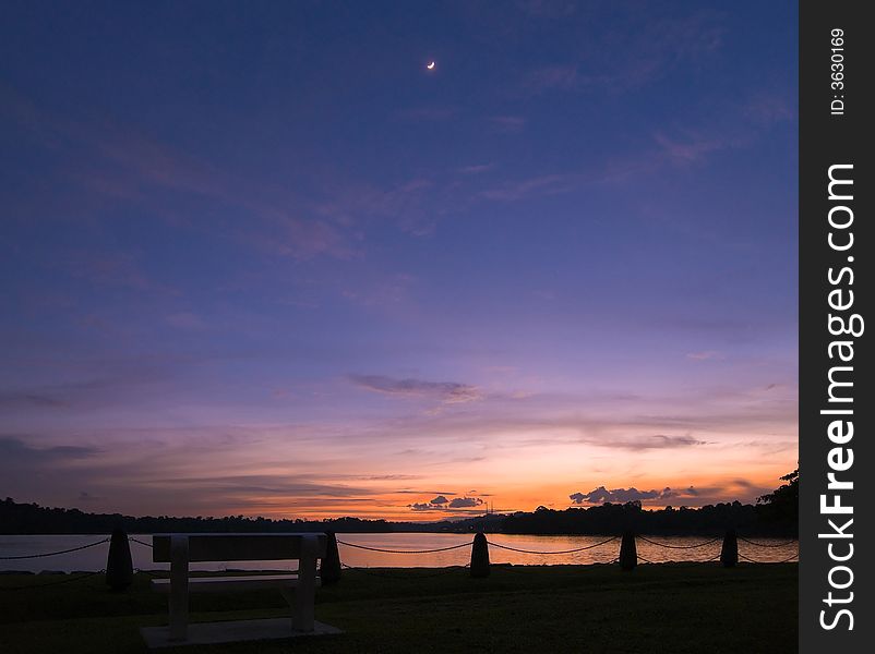 A bright crescent moon high in the twilight sky above an empty stone bench in a public park \. A bright crescent moon high in the twilight sky above an empty stone bench in a public park \