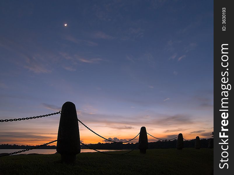 A bright crescent moon high in the twilight sky framed by a chain fence on the shores of a lake. A bright crescent moon high in the twilight sky framed by a chain fence on the shores of a lake