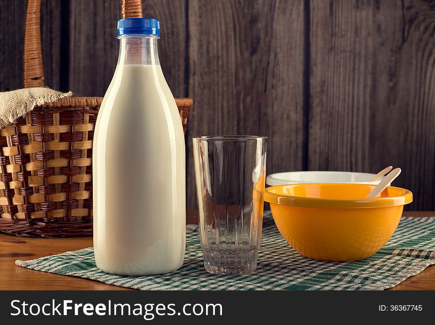 Still life of plastic bottle of milk, empty glass, two plastic bowls and wicker basket over checked table-napkin. Wooden wall background. Still life of plastic bottle of milk, empty glass, two plastic bowls and wicker basket over checked table-napkin. Wooden wall background.