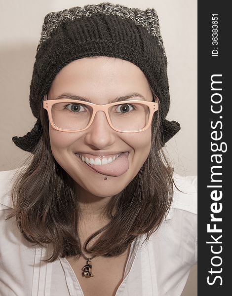 Close-up portrait of young cheerful girl showing her tongue. She is wearing funny black color winter hat, pinky glasses and white shirt. Isolated. Close-up portrait of young cheerful girl showing her tongue. She is wearing funny black color winter hat, pinky glasses and white shirt. Isolated
