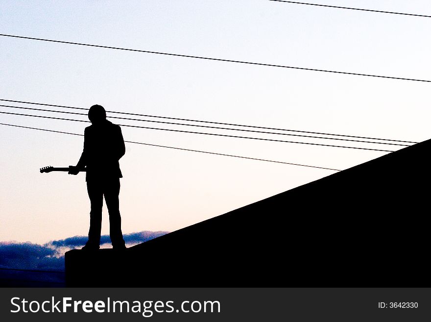A musician and his silhouette are photographed at dusk with power lines and clouds in the background of the sunset sky. A musician and his silhouette are photographed at dusk with power lines and clouds in the background of the sunset sky.