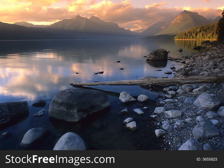 A calm mountain lake and shoreline with clouds and mountains in the background.