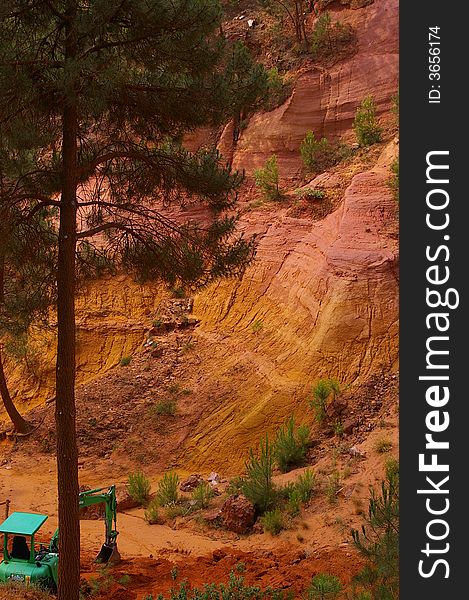 Geological landscape composed by a red and yellow rock sculpted by erosion and pine wood. 
There is a green crane.
It is in the south of France, a place called Luberon. Geological landscape composed by a red and yellow rock sculpted by erosion and pine wood. 
There is a green crane.
It is in the south of France, a place called Luberon.
