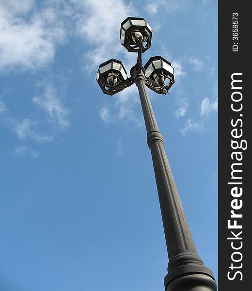 Black vintage street lamp pole, with 3 bulbs, against blue, slightly cloudy, sky.