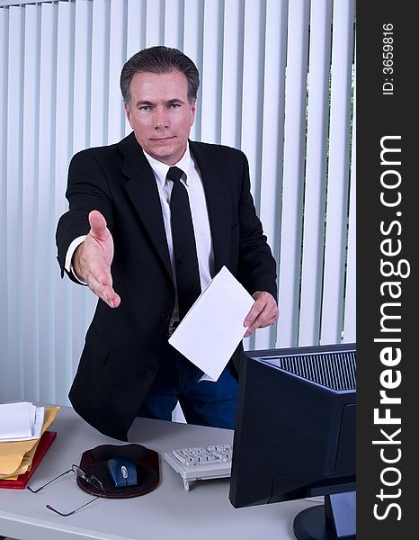 A man standing in front of a desk with his hand extended as if to greet someone. A man standing in front of a desk with his hand extended as if to greet someone.