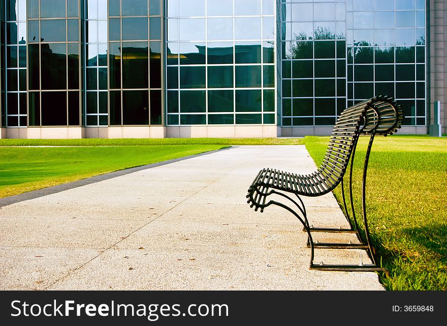 An empty park bench outside of a large glass paned building. An empty park bench outside of a large glass paned building.