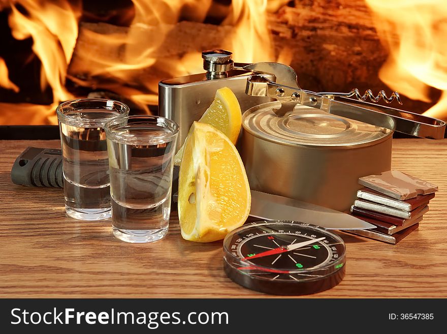 Two glasses of alcohol, knife, compass, canned meat, chocolate, and hip flask with open fire. Two glasses of alcohol, knife, compass, canned meat, chocolate, and hip flask with open fire
