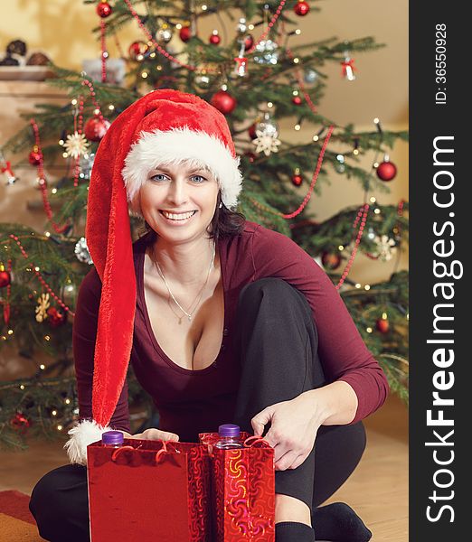 Beautiful middle age woman in red party santa hat sitting near the Christmas tree with gifts. Beautiful middle age woman in red party santa hat sitting near the Christmas tree with gifts