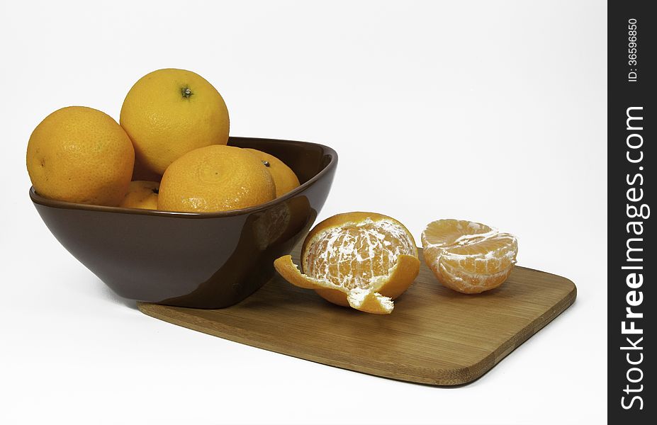 Clementine oranges in a brown bowl and unpeeled on a bamboo cutting board against a white background. Clementine oranges in a brown bowl and unpeeled on a bamboo cutting board against a white background