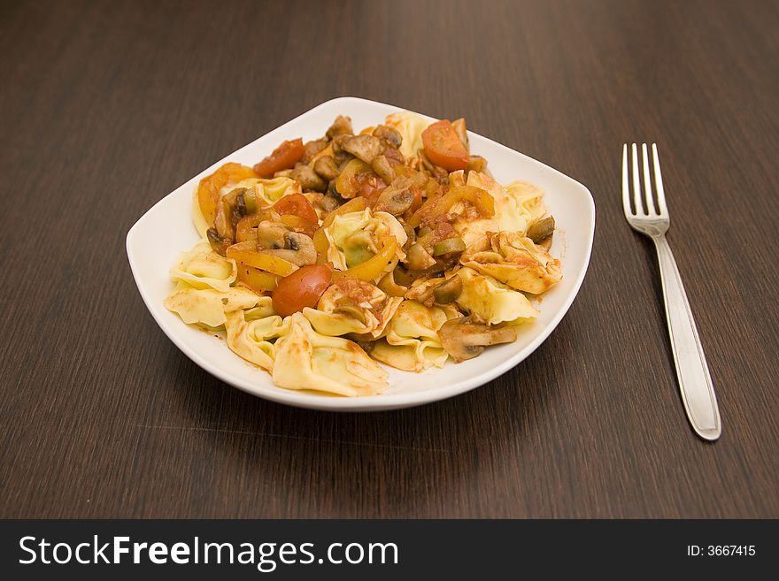 Tortellini with vegetables (mushrooms, peppers, tomatoes) in a plate on a wenge table and a fork