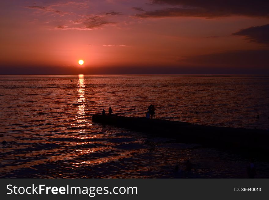 The photo shows a sunset over the sea and the silhouettes of people standing on the coast. The photo shows a sunset over the sea and the silhouettes of people standing on the coast.