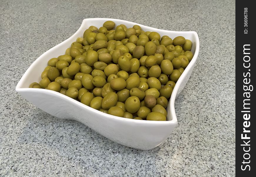 Aerial view of a green olives contained in a white square bowl resting on a kitchen counter top. Aerial view of a green olives contained in a white square bowl resting on a kitchen counter top