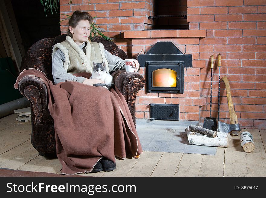 Mature woman sitting with cat in armchair near stove in country house. Mature woman sitting with cat in armchair near stove in country house