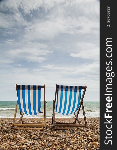 Portrait shot of 2 stripy blue and white deck chairs on a shingle beach in England looking out to sea. Portrait shot of 2 stripy blue and white deck chairs on a shingle beach in England looking out to sea