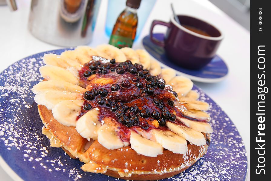 A gourmet waffle topped with sugar, banana and berries, set on a white table with coffee & syrup. A gourmet waffle topped with sugar, banana and berries, set on a white table with coffee & syrup