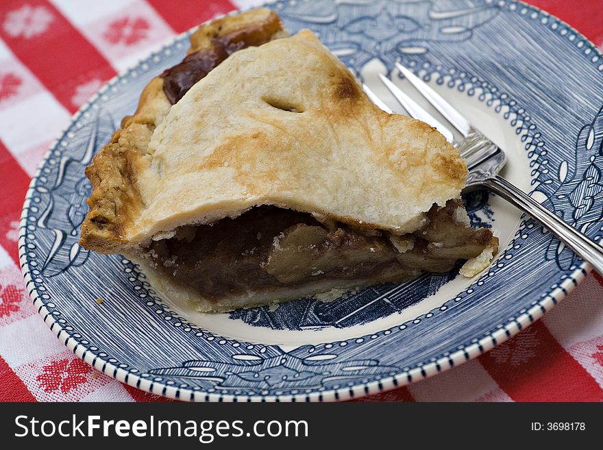 A slice of apple pie on old blue and white plate, fork and red and white gingham tablecloth